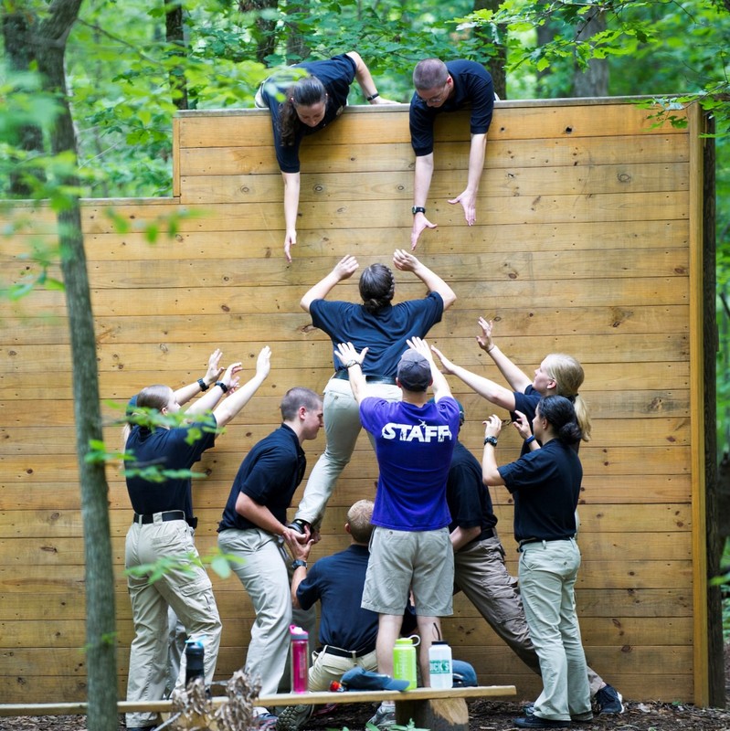 A team of people on an obstacle course work together to boost one member of the group over a high wall. A team of people on an obstacle course work together to boost one member of the group over a high wall.