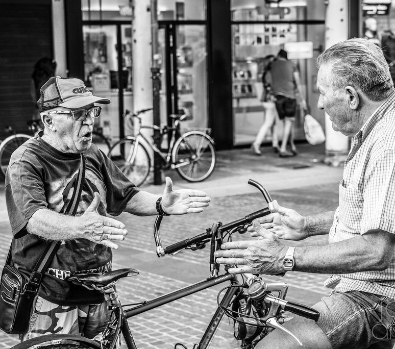 Two men stand talking in the street, each making nearly identical hand gestures. Two men stand talking in the street, each making nearly identical hand gestures.