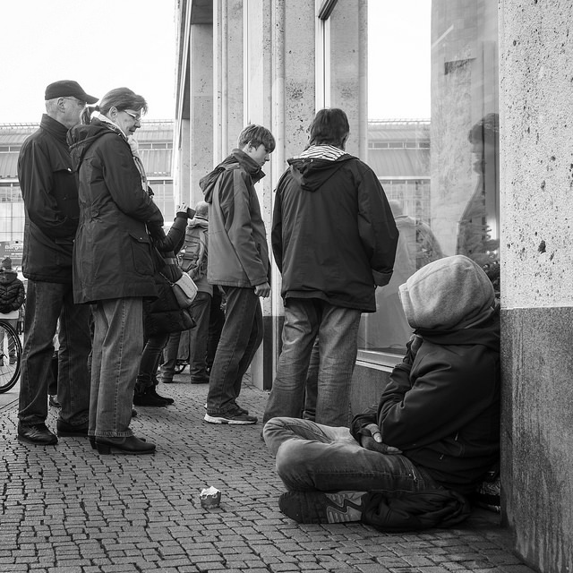 A homeless man sits on the sidewalk on a busy street as crowds of shoppers stand nearby. A homeless man sits on the sidewalk on a busy street as crowds of shoppers stand nearby.